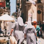 Two nuns in grey habits walk down a sunlit street, each carrying shopping bags. The street is bustling with people. Old buildings line the pathway, and a canopy-covered outdoor seating area is visible in the background. The scene exudes a mix of modern and historic charm.