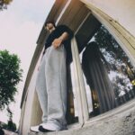 A person wearing a black shirt, grey sweatpants, and white sneakers stands on the edge of a window ledge of a building. The camera angle is from below, creating a dramatic perspective. Trees and part of the street are visible in the background.