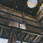 A person stands on a platform inside a tall, industrial building structure with large circular openings in the roof, allowing natural light to filter through. The walls and ceiling are made of metal and beams, creating a complex, geometric pattern.