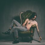 A shirtless person with tattoos is lounging on a vintage, green, high-back armchair. They are wearing plaid pants and black shoes with socks. The person is posed with one leg draped over the arm of the chair and one arm hanging down, creating a relaxed, contemplative mood.