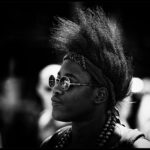 A black-and-white photograph features a person with a tall, styled mohawk and wearing round sunglasses. The individual has a bandana tied around their head and a beaded necklace. The background is blurred, highlighting the person as the focal point of the image.