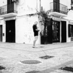 A man stands on an empty cobblestone street corner in a European town. He wears shorts and a t-shirt and uses a phone. Buildings with shuttered windows, balcony railings, and lampposts surround him. The scene is in black and white.