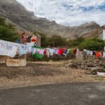 Clothes are hung to dry on a line strung between stone buildings and trees in a rugged, mountainous area. The landscape is arid with sparse vegetation, and the sky above is partly cloudy. One of the buildings is white and situated near rocky terrain.