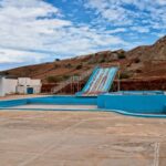 An empty, abandoned outdoor water park with blue slides and pools set against a barren, rocky hillside. The pools are dry, and the surrounding structures are weathered. The sky above is partly cloudy.