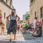 A group of people walks down a narrow, cobblestone street flanked by historic stone buildings. Tourists are dressed in summer clothes, with one woman in a colorful skirt standing with hands on hips. A child in a hat sits on the side, observing the crowd.