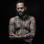 A man with a full beard and glasses stands against a black backdrop. He is shirtless, showcasing extensive tattoos that cover his arms, chest, and neck. He is posed with his arms crossed, displaying intricate and detailed tattoo designs.