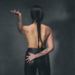 A person, viewed from behind, stands against a dark background. They have long, braided hair and tattoos on their arms. They are shirtless and wear black pants, with one arm raised and the other resting on their lower back.