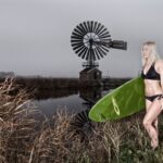 A woman in a black bikini stands by a grassy riverside, holding a green surfboard under her arm. Behind her is a calm river and a rustic windmill structure against a cloudy, overcast sky.