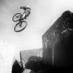 A cyclist is captured in mid-air while performing a jump over large, angular rocks. The image is in black and white, emphasizing the contrast and dramatic action against the bright sky. The rider is wearing a helmet and appears to be engaged in an extreme biking stunt.