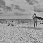 A black-and-white photo of a beach with a few people. In the foreground, a person stands with a blanket or towel blowing in the wind. In the background, another person sits on a towel, and two more individuals are near the shoreline with gentle waves and clouds above.