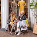 Two women are sitting on a bench outside a clothing store with mannequins in the window. They are laughing and holding shopping bags, one with a yellow scarf and showcasing a stylish outfit. The setting is on a brick-paved street with a small tree nearby.