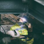 A person in safety gear, including a helmet and high-visibility jacket, is welding a metal structure. The welding torch emits a bright light, and the scene appears to be part of a construction or maintenance project. The person is focused on their work amidst steel beams.