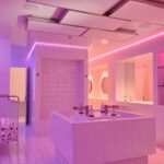 A modern, neon-lit bathroom with white tiled walls and floors. The room features a large central sink island with dual faucets, a rack holding toilet paper rolls, and a wall-mounted mirror with shelves beneath it. The neon lights cast a pink and purple hue.