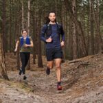 A man and a woman run on a rugged forest trail. The man, in the foreground, wears a backpack, shorts, and a long-sleeve shirt. The woman, slightly behind, wears a tank top and shorts. Both seem focused and energized, running among tall trees on an uneven path.