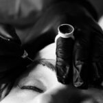 Close-up black and white image of a person undergoing a microblading procedure. A technician wearing black gloves is using a tool to apply pigments to the person's eyebrow. A ring with a small container of pigment is visible on one of the technician's fingers.