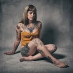 A person with various tattoos on their arms, legs, and torso sits on the floor against a gray concrete wall. They have long hair with bangs, wear a sleeveless yellow top, and gaze calmly into the camera.