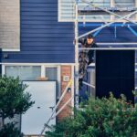 A construction worker wearing a harness climbs a metal scaffold in front of a blue, two-story house. Green shrubs are in the foreground, and building materials are scattered around the area. The worker appears to be focused on the task at hand.