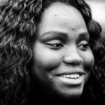 Black and white close-up photo of a person with long, curly hair, displaying a candid smile and looking slightly off to the side. Their expression appears content and thoughtful. The background is blurred.