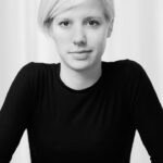 Black and white portrait of a person with short, light-colored hair, wearing a long-sleeved black shirt. They are looking directly at the camera with a neutral expression, set against a plain, light background.