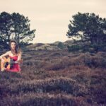 A woman in a red dress sits among shrubs in a field, playing an acoustic guitar. Two large trees stand in the background under an overcast sky. The scene has a natural, serene atmosphere.