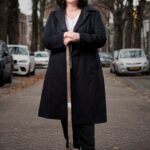 A person with curly hair and glasses is standing on a tree-lined street holding a shovel. They are wearing a dark overcoat and casual clothing. Cars are parked on both sides of the street, and autumn leaves are scattered on the ground.