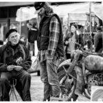 Black and white image of an outdoor market. An elderly man with a beard, wearing a beret, sits on a stool holding a card. Beside him, a long-haired man in a cap and plaid shirt stands. In the background, people and various items, including vintage motorcycles, are visible.