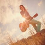 A person stands in a field with tall grass, holding an acoustic guitar. The sun shines brightly behind them, creating a lens flare effect. The sky is partly cloudy, with a mixture of blue and white hues. The person wears a dark top and light-colored pants.