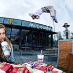 A young woman drinks from a McDonald's cup and holds fries while sitting at an outdoor table laden with McDonald's food. Behind her, a person performs a mid-air flip in front of the McDonald's building, with a few onlookers in the background.