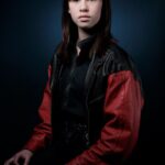 A young person with shoulder-length dark hair sits against a dark backdrop. They are wearing a black and red leather jacket over dark clothes, looking directly at the camera with a neutral expression.