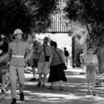 A black and white photo of a man in a historical military uniform walking down a tree-lined path. Tourists, some with cameras and maps, explore the area. Tall trees frame the walkway, and sunlight creates dappled shadows on the ground.