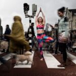 A woman in colorful athletic wear stands on one leg in a yoga pose at a crosswalk, surrounded by blurred pedestrians and a small dog. The setting appears urban, with buildings and a traffic light in the background. The scene conveys a sense of calm amidst chaos.