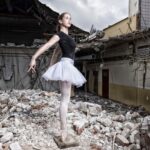 A ballerina in a black top and white tutu poses gracefully on her toes amid the ruins of a dilapidated building, surrounded by rubble and debris. The collapsed structure forms a stark contrast to her elegant stance, symbolizing resilience and hope.