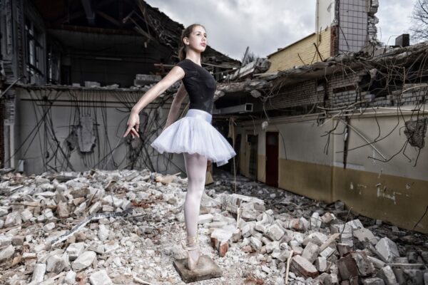 A ballerina in a black top and white tutu poses gracefully on her toes amid the ruins of a dilapidated building, surrounded by rubble and debris. The collapsed structure forms a stark contrast to her elegant stance, symbolizing resilience and hope.