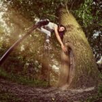 A person with long hair and wearing athletic clothes uses stilts to reach and touch a large tree in a lush forest. The person appears to be performing an acrobatic stunt, with one leg on the ground and the other elevated.