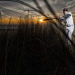 A baseball player in uniform poses as if swinging a bat in a field of tall grass. The sky is dramatic with streaks of clouds and the setting sun creates a striking backdrop, illuminating the scene with warm, golden light.