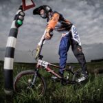 A BMX rider, dressed in orange and blue racing gear and a black helmet, waits at a traffic light on a grassy hill under a cloudy sky. The bike has the number 84 on the front. The background features an open field and a line of trees in the distance.