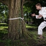 A martial artist in a white gi stands in a defensive pose in front of a large tree. The tree has a white belt tied around its trunk. The scene is set in a lush, green park with dappled sunlight filtering through the leaves.