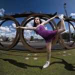 A young girl in a purple dress and ice skates balances on one leg with the other leg extended behind her. She poses on a green surface with large circular metal sculptures and a blue sky with clouds in the background.