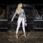 A woman dressed in white riding attire and red shoes stands confidently in front of two horse stalls. She holds a riding crop and a top hat, with a stable and two horses visible behind her. The scene is lit dramatically, emphasizing her stance and the surroundings.