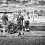 A black and white photo features three street cleaners standing on a sidewalk. The person in the center holds a broom and looks directly at the camera, while the other two engage in tasks with their cleaning cart. Cars and buildings are visible in the background.