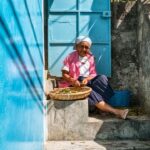 An elderly woman, wearing a white headscarf and a pink shirt, sits on stone steps next to a blue gate. She is sorting through a basket of produce in her lap, with a blue wall beside her. Sunlight casts shadows over the scene, highlighting the textures and colors.