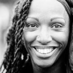 Black and white close-up of a person smiling warmly at the camera. They have braided hair partially covered by a headscarf and a small nose piercing. The background is blurred, drawing focus to their face.