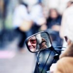 Close-up of a person with glasses and white hair, seen in a side mirror of their mobility scooter. The person wears a tan coat and the background is blurred, showing indistinct figures and an outdoor environment, suggesting a busy street or market scene.