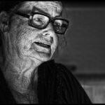 Black and white close-up of an elderly person with glasses and deep wrinkles, looking slightly away from the camera. The lighting highlights the texture of their skin, creating a dramatic and evocative image.