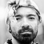 A monochrome close-up of a man with a beard and mustache, gazing intently at the camera. He is wearing a patterned cloth wrapped around his head like a turban. The background is blurred, focusing attention on his facial expression.