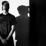 A man stands with his hands clasped in front of him, wearing a T-shirt with an intricate design and a cap that reads "Death". He is leaning against a wall, casting a distinct shadow. The image is in black and white, creating a dramatic contrast.