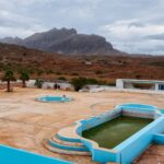 A deserted swimming pool area with blue-painted pools, surrounded by an arid, rocky landscape. Palm trees and a few small buildings are present, with mountains in the background under a cloudy sky. The scene appears quiet and abandoned.