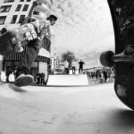 Black and white photo of skateboarders in an urban setting. One skater is about to perform a trick off a ramp, while another observes closely. Several people in the background watch near modern buildings and a cloudy sky. Two skateboards are visible in the foreground.