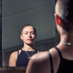 A person with a shaved head and wearing a black sleeveless top looks at their reflection in a mirror. The room is softly lit, and the background is blurred, bringing focus to the person's expression and the mirror's reflection.