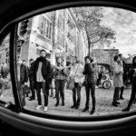 Black and white photo of a group of people standing on a city street, viewed through the windshield of a car. Some individuals are using their phones, others are observing their surroundings. The background features a large building and trees.